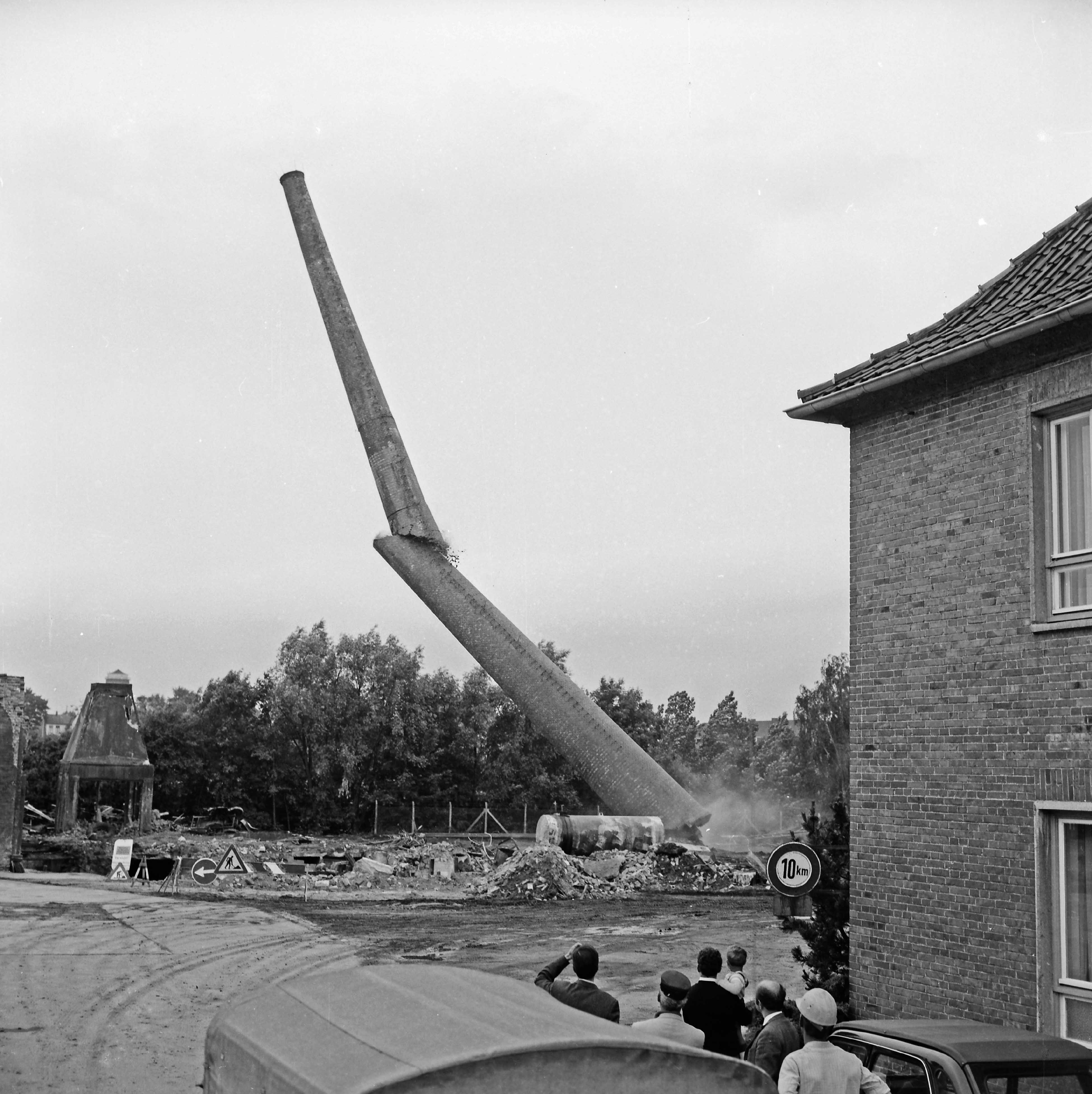 Bad Oldesloe: Stadtwerke Bad Oldesloe: Gaswerk: Lübecker Straße: Sprengung des Schornsteins des abgerissenen Kraftwerks: rechts Gebäudeecke: vorn Wagendach und Zuschauer: dahinter stark geneigter, in der Mitte gebrochener Schornstein mit Gastank, am Fuß Rauchwolken: hinten Bauschutt: links teilweise abgerissenes Gebäude: hinten Bäume: 10. Juni 1971