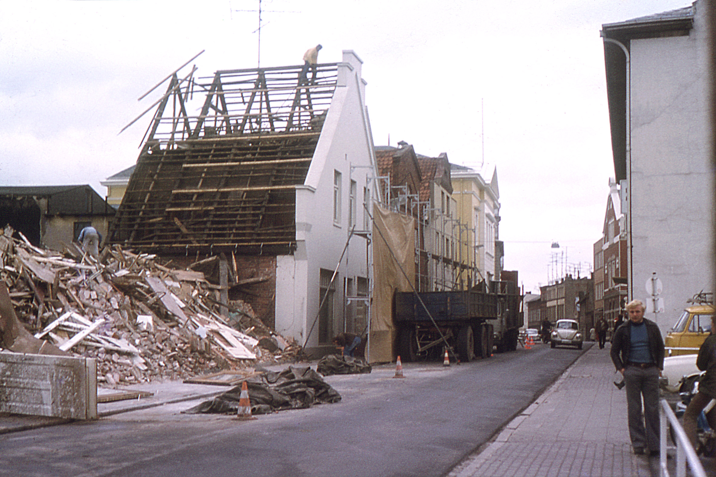 Bad Oldesloe: Hagenstraße: Abriss von Gebäuden für Neubau Kreissparkasse Stormarn: links Bauschutthaufen, dahinter Haus ohne Dach: darauf Arbeiter: davor Absperrung und Lkw: vorn rechts Mann: dahinter Hauptgebäude der Kreissparkasse, Stadthaus und Pkw: hinten Mitte Rathaus, 1973