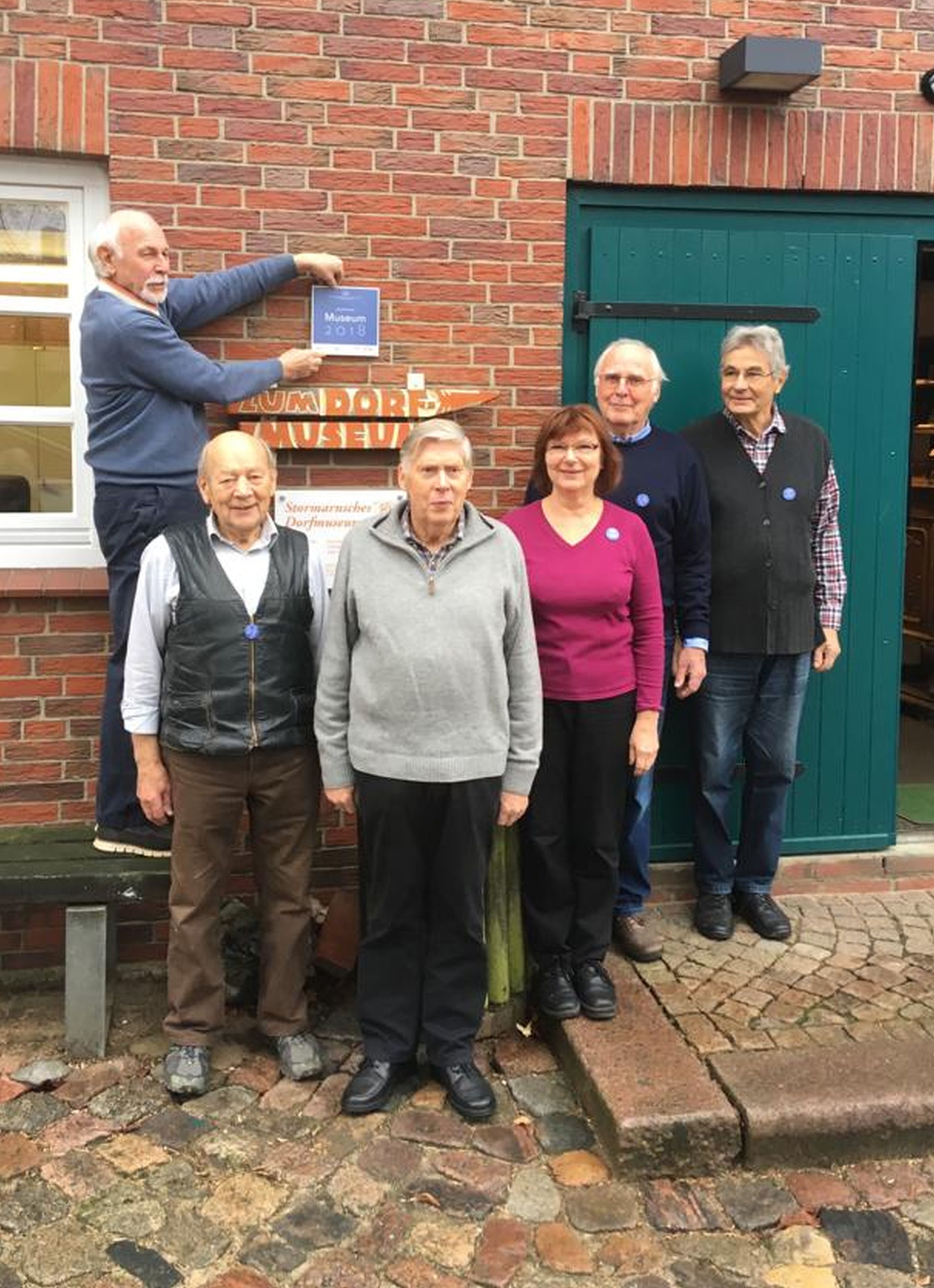 Eine kleine Gruppe der ehrenamtlichen Mitarbeiter des Museums, die erfolgreich die Zertifizierung abgeschlossen haben, zeigen stolz ihre Plakette des Landes Schleswig-Holstein am Dorfmuseum. 